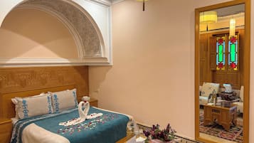 A hotel room with a large bed, a wooden headboard, a small table with a vase of flowers, and a mirror reflecting a door with stained glass.