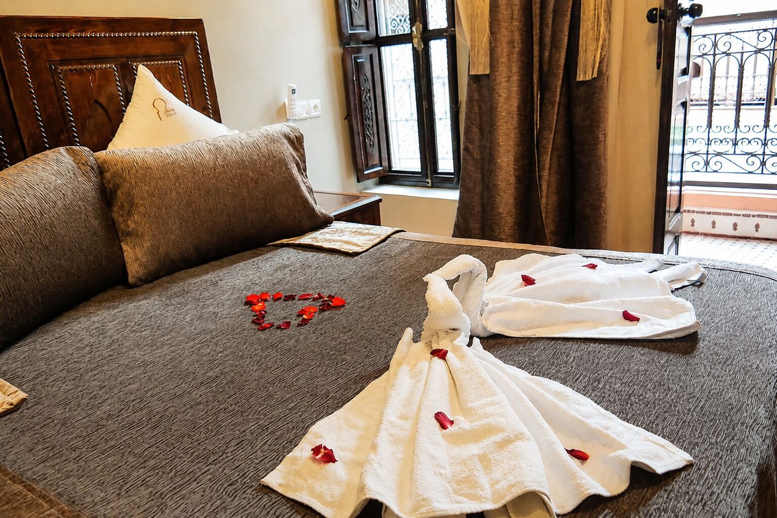 A bed with a brown textured bedspread, two white towels folded on it, and red rose petals arranged on the bed.