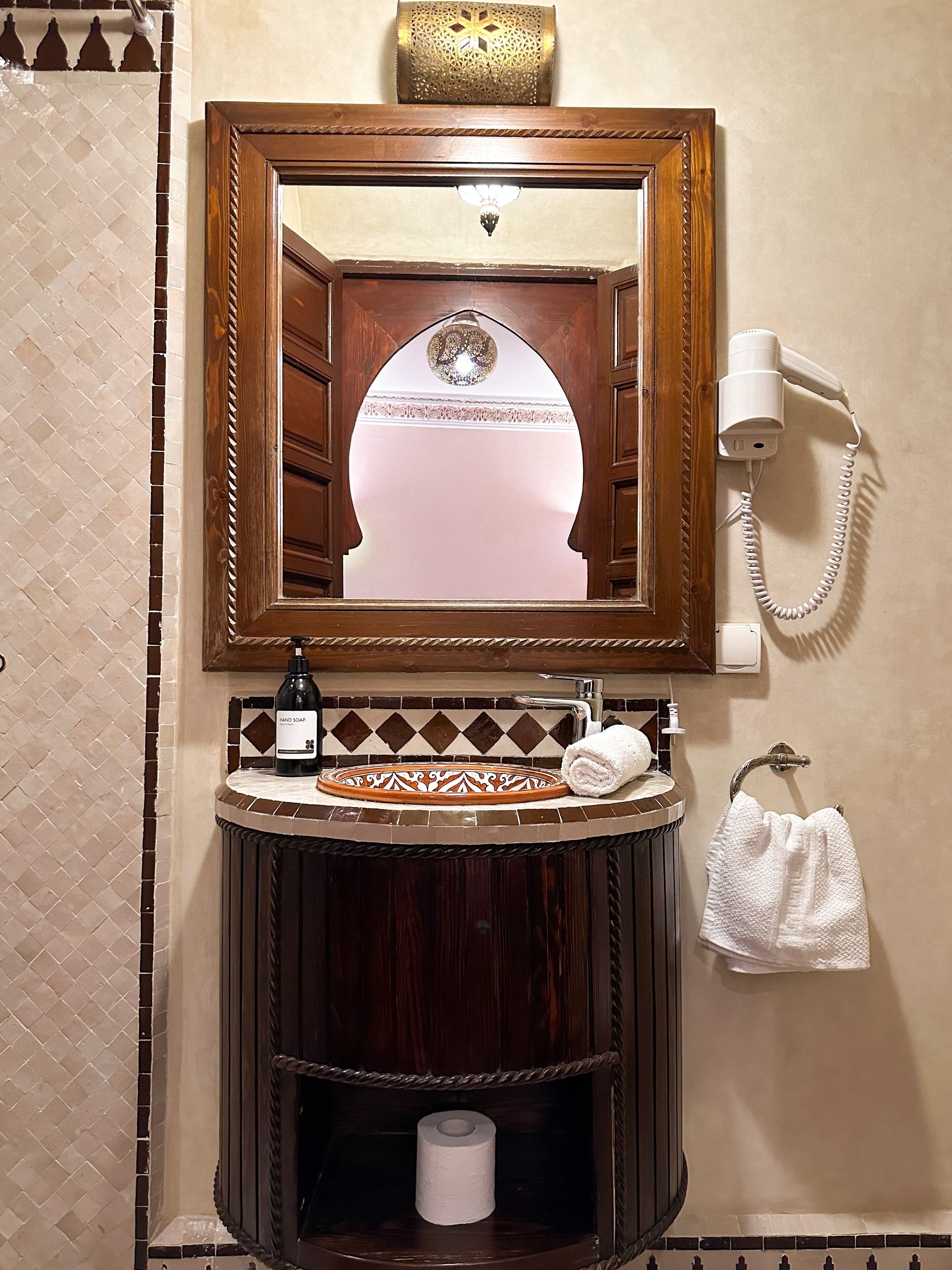 A bathroom with a wooden-framed mirror, a hand soap dispenser, a towel, and a hairdryer on the wall.