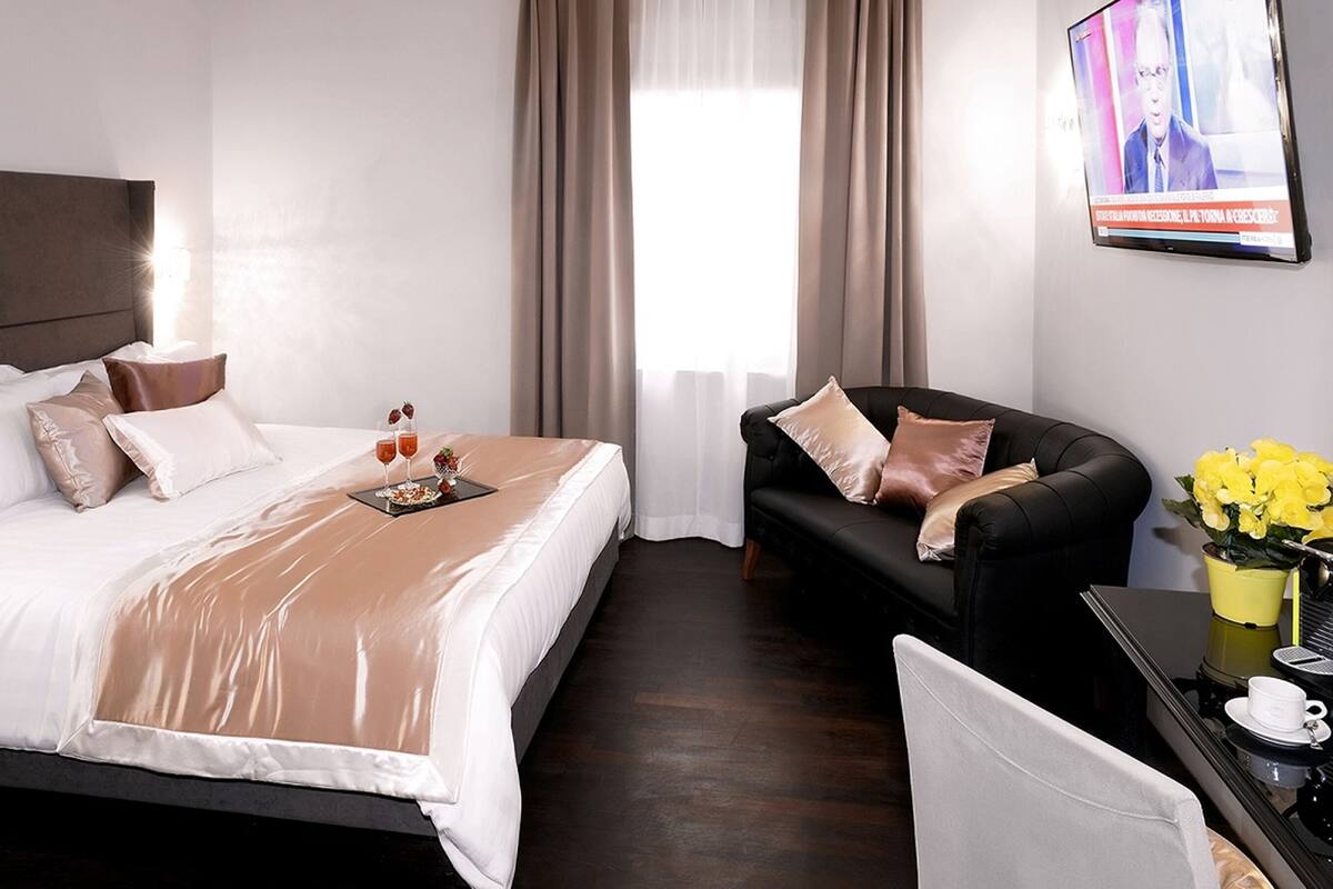 Habitación de hotel con cama, sofá, escritorio con cafetera y televisor en la pared.