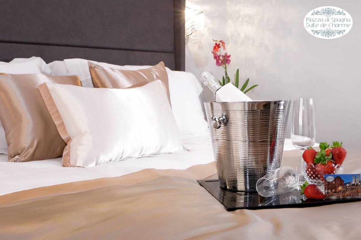 Una cama bien hecha con almohadas blancas y color beige, un cubo metálico con una flor, y una bandeja con fresas y una postal.