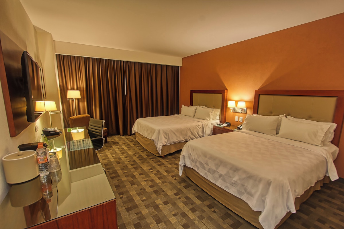 Une chambre d’hôtel avec deux lits, un bureau avec un miroir, une chaise, une lampe et une grande fenêtre avec des rideaux.