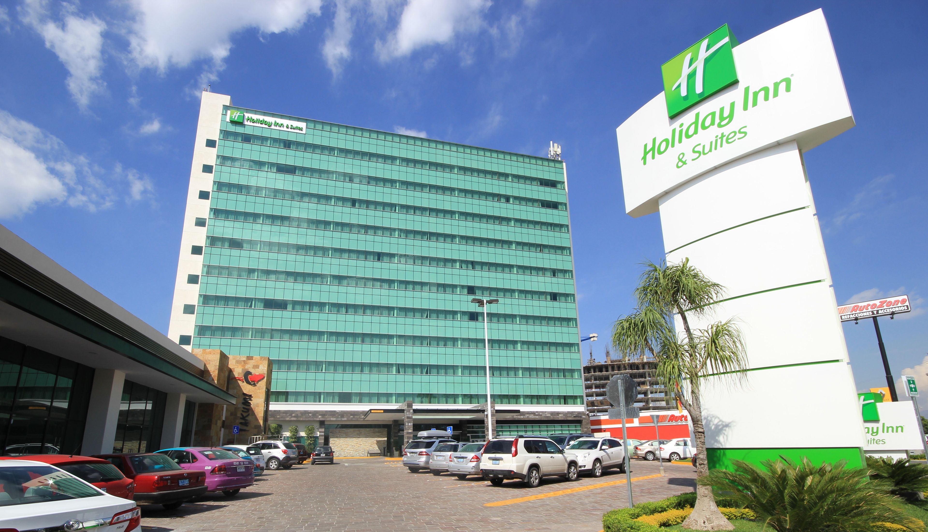 Edificio de Holiday Inn & Suites con fachada de vidrio moderna, estacionamiento con varios coches y un letrero de AutoZone cerca.