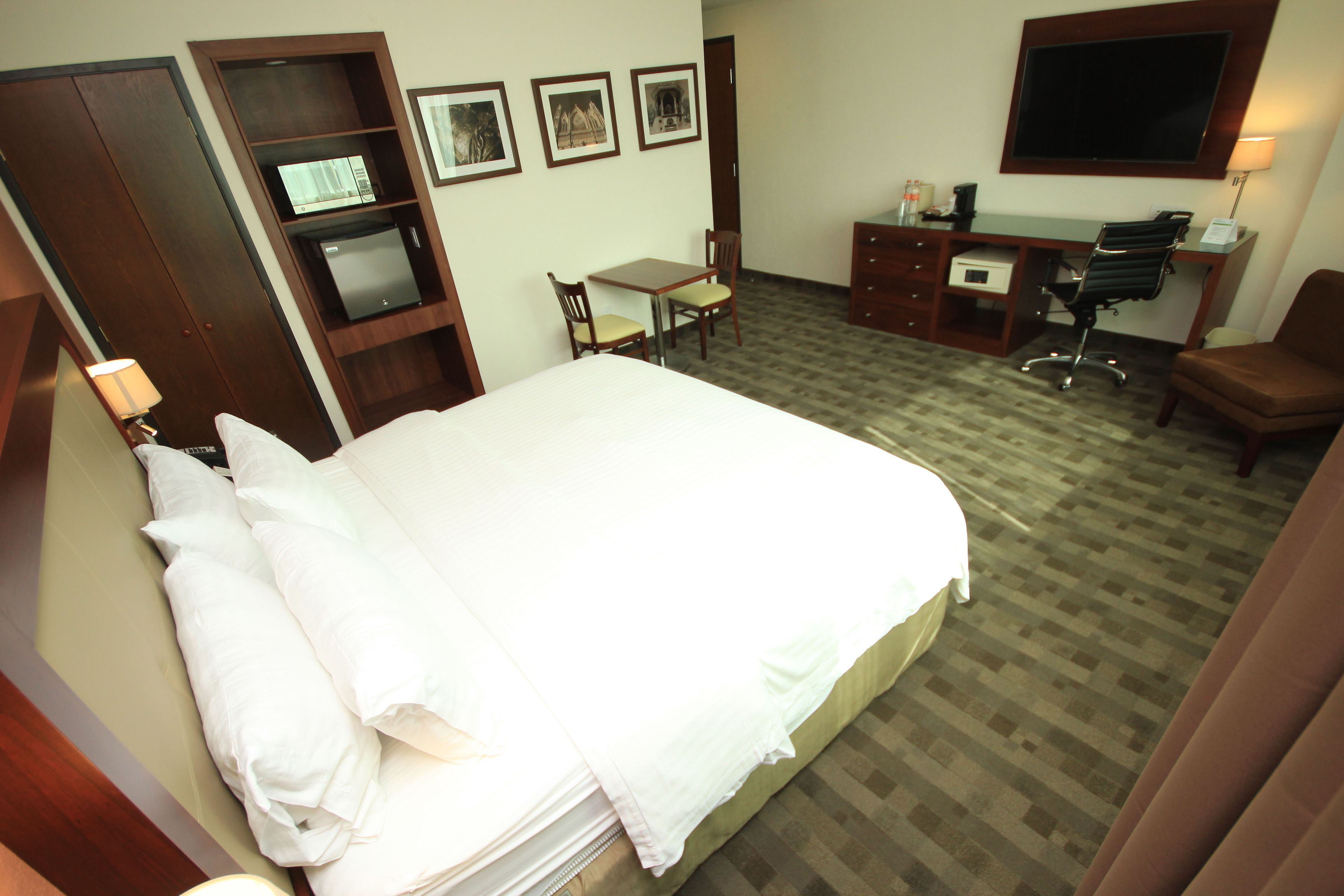 Quarto de hotel com cama, escrivaninha, cadeira, televisão e guarda-roupa.