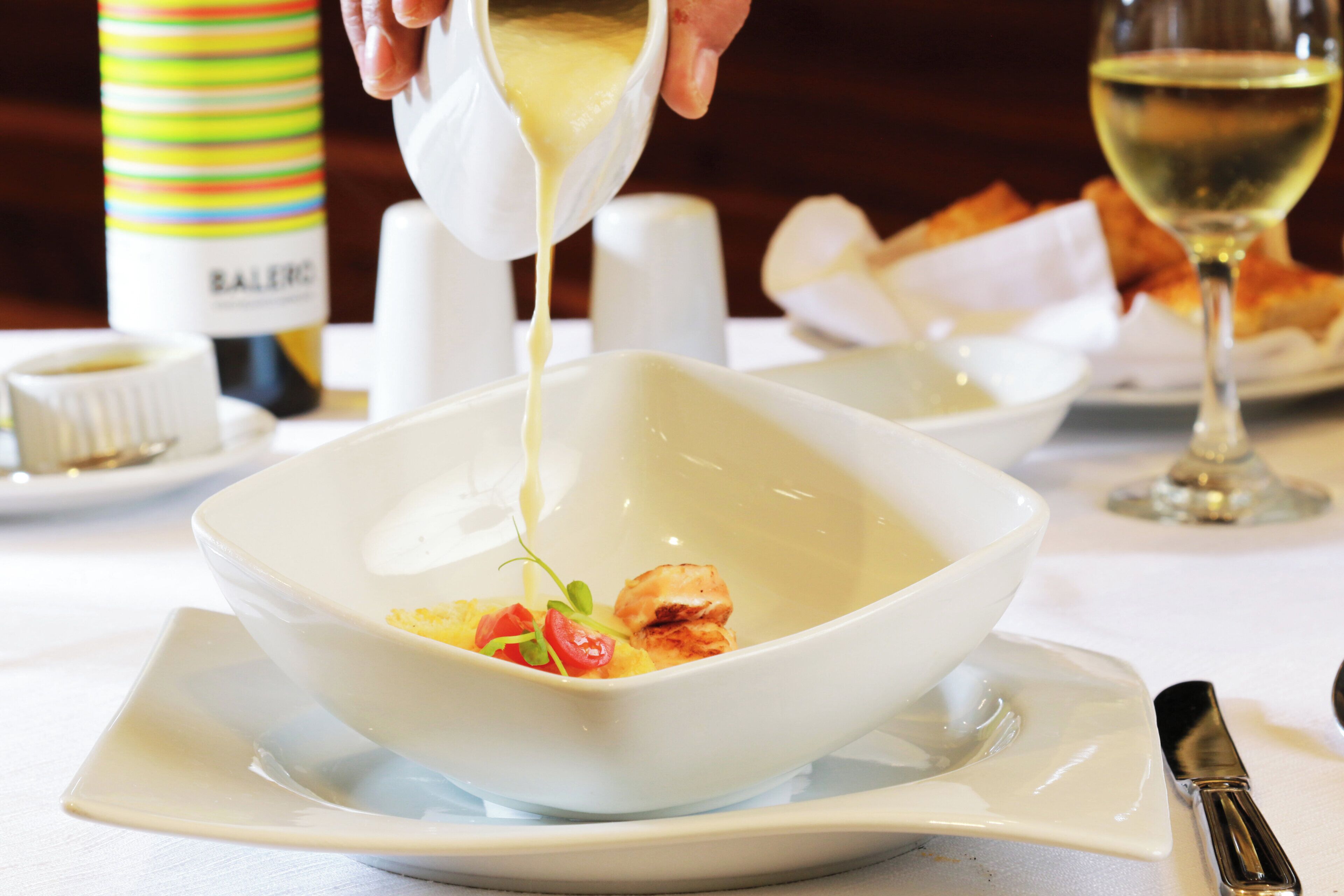 A white bowl on a plate with a sauce being poured into it, a bottle of sauce, a glass of white wine, and a napkin on the table.
