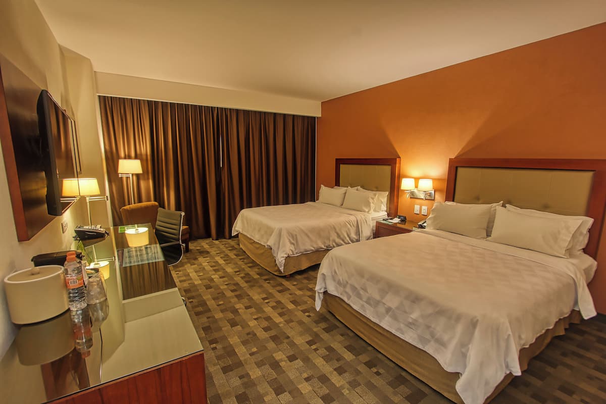 A hotel room with two beds, a desk with a mirror, a chair, a lamp, and a large window with curtains.