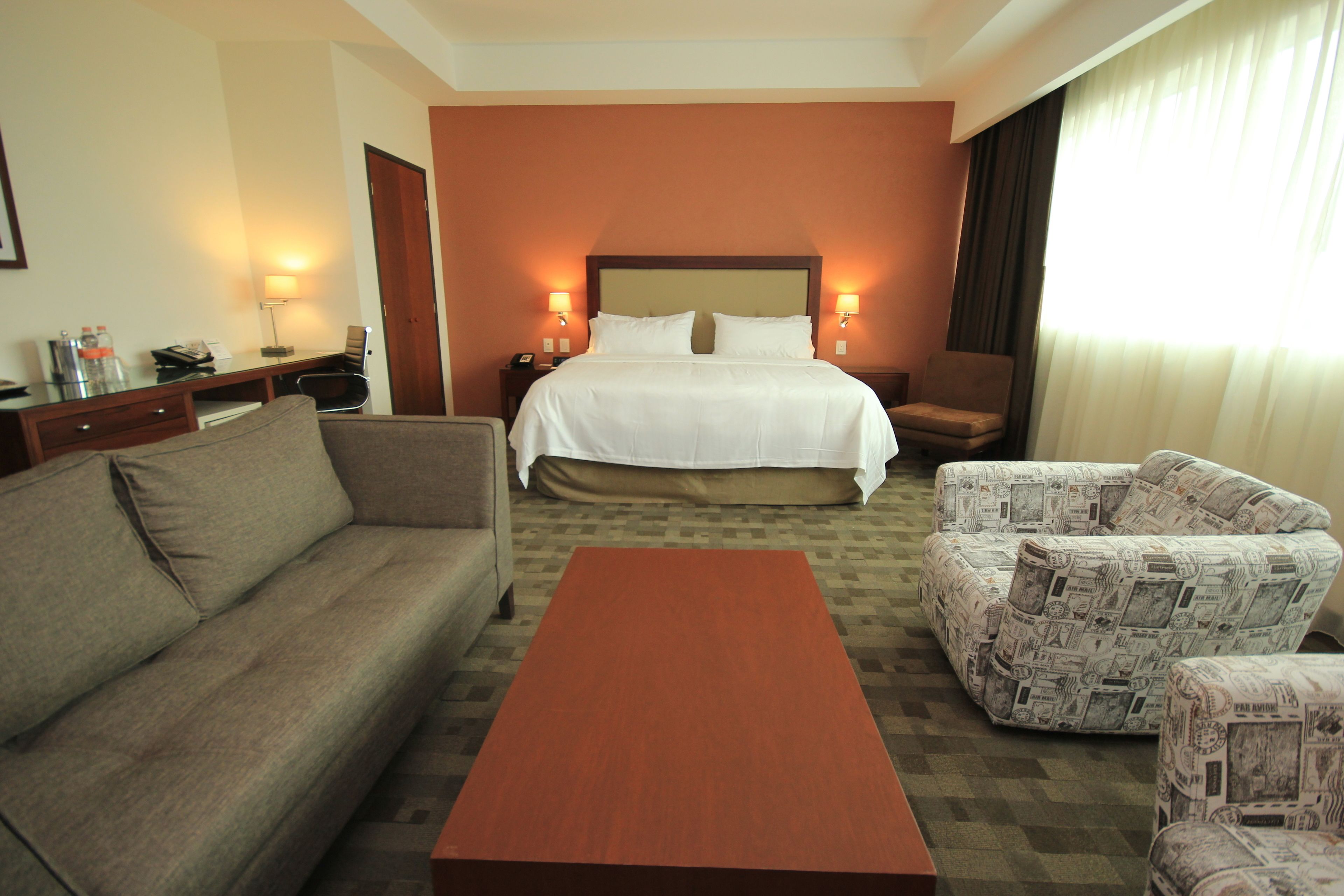 A hotel room with a bed, a sofa, two armchairs, a wooden coffee table, and a desk with a lamp.
