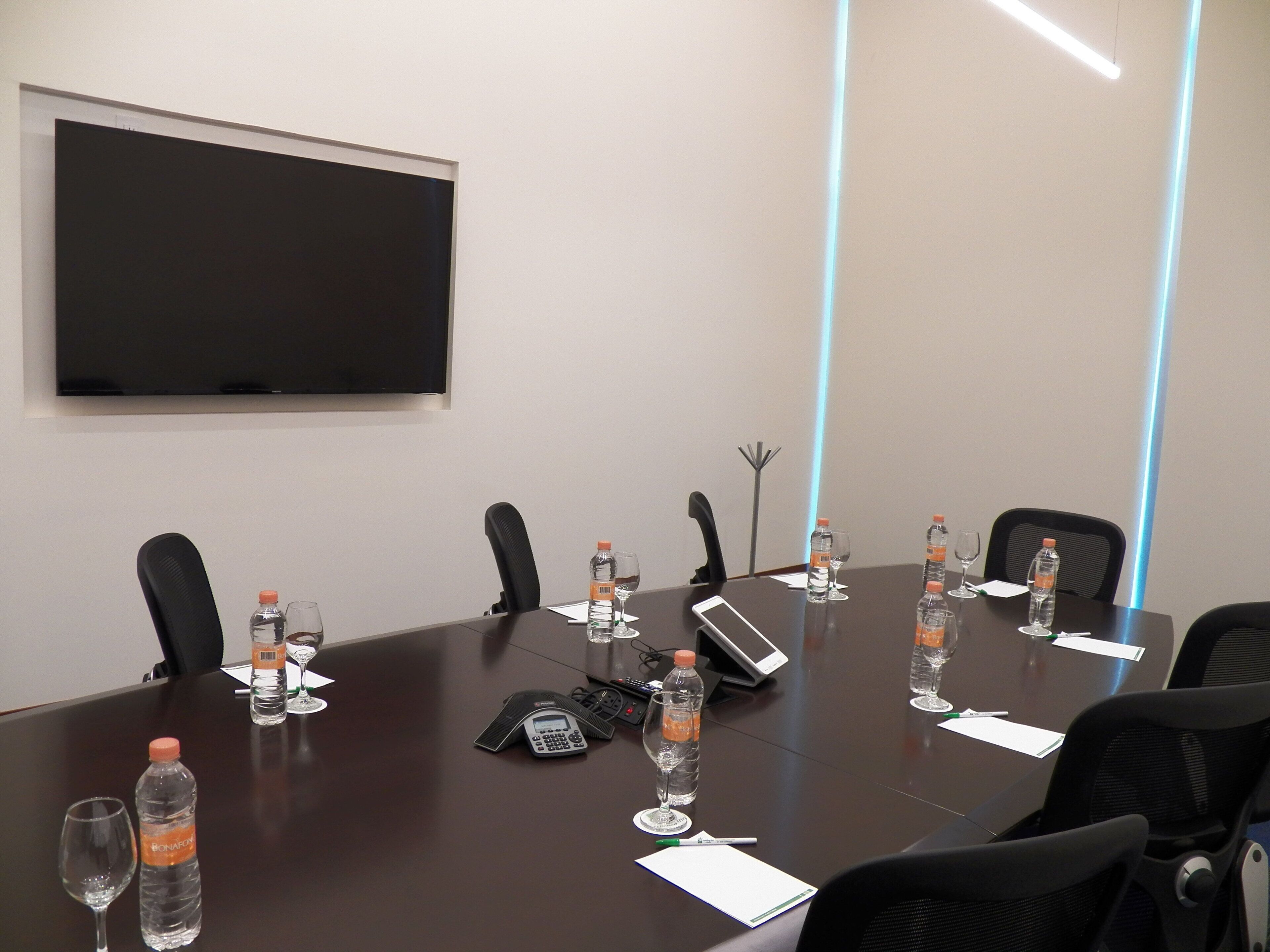 Un salón de conferencias con una gran mesa, sillas, una pantalla de proyector, botellas de agua, vasos y un teléfono.