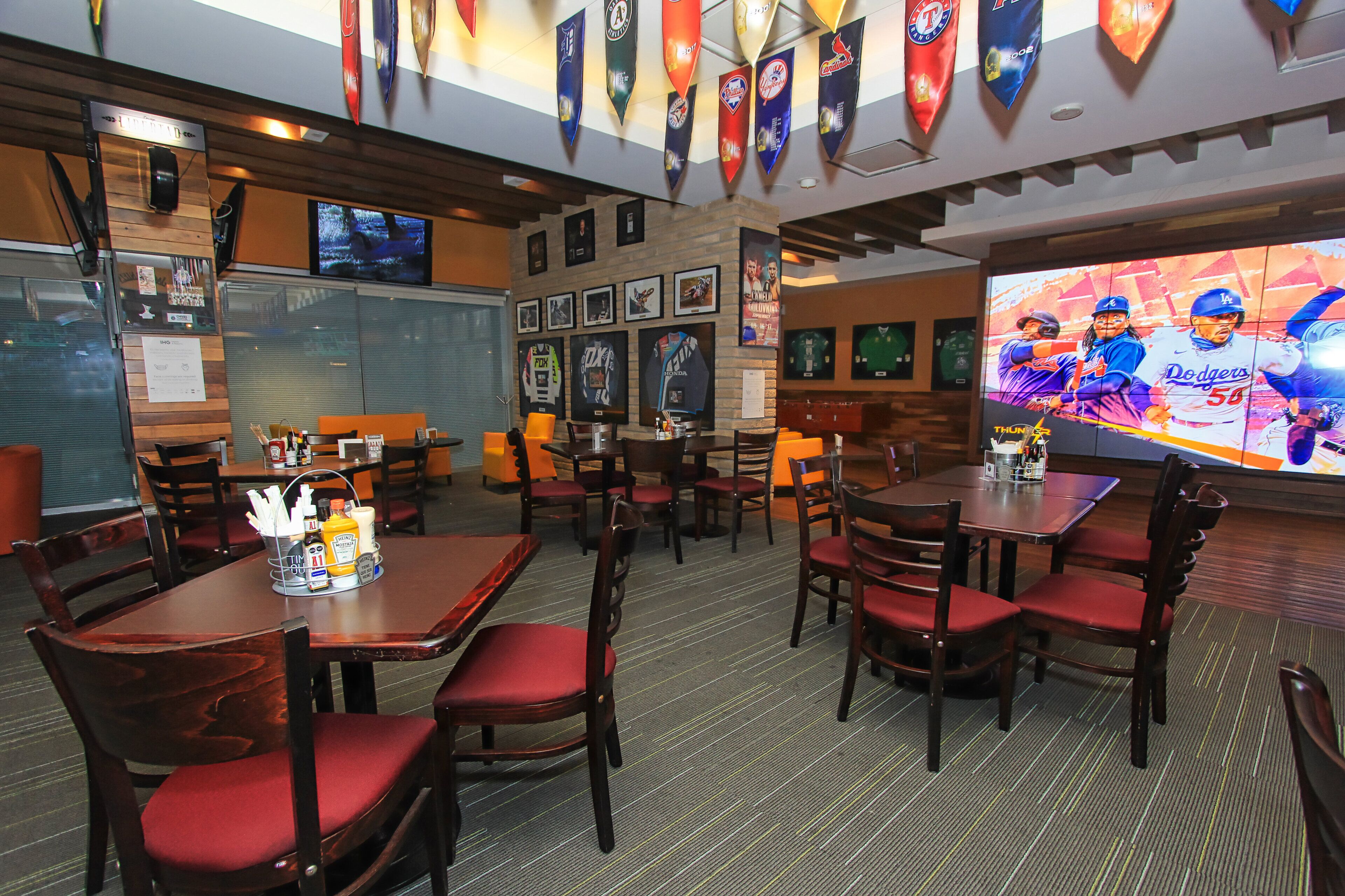 Restaurante com mesas de madeira e cadeiras vermelhas estofadas, uma grande tela de TV exibindo esportes e diversas bandeiras esportivas no teto.