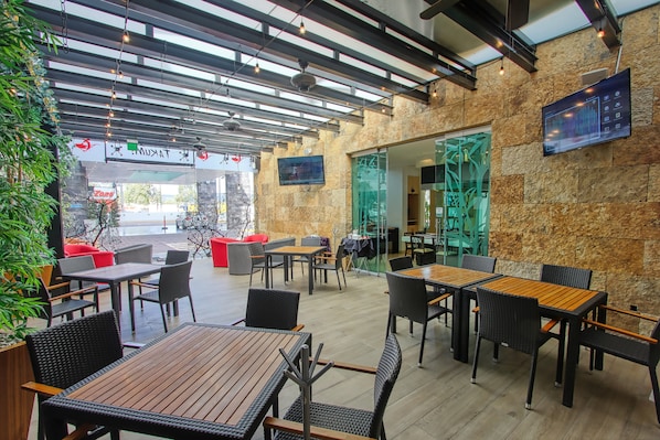 Un espace café en plein air avec des tables et des chaises, un mur en pierre et une porte vitrée donnant sur l’intérieur.