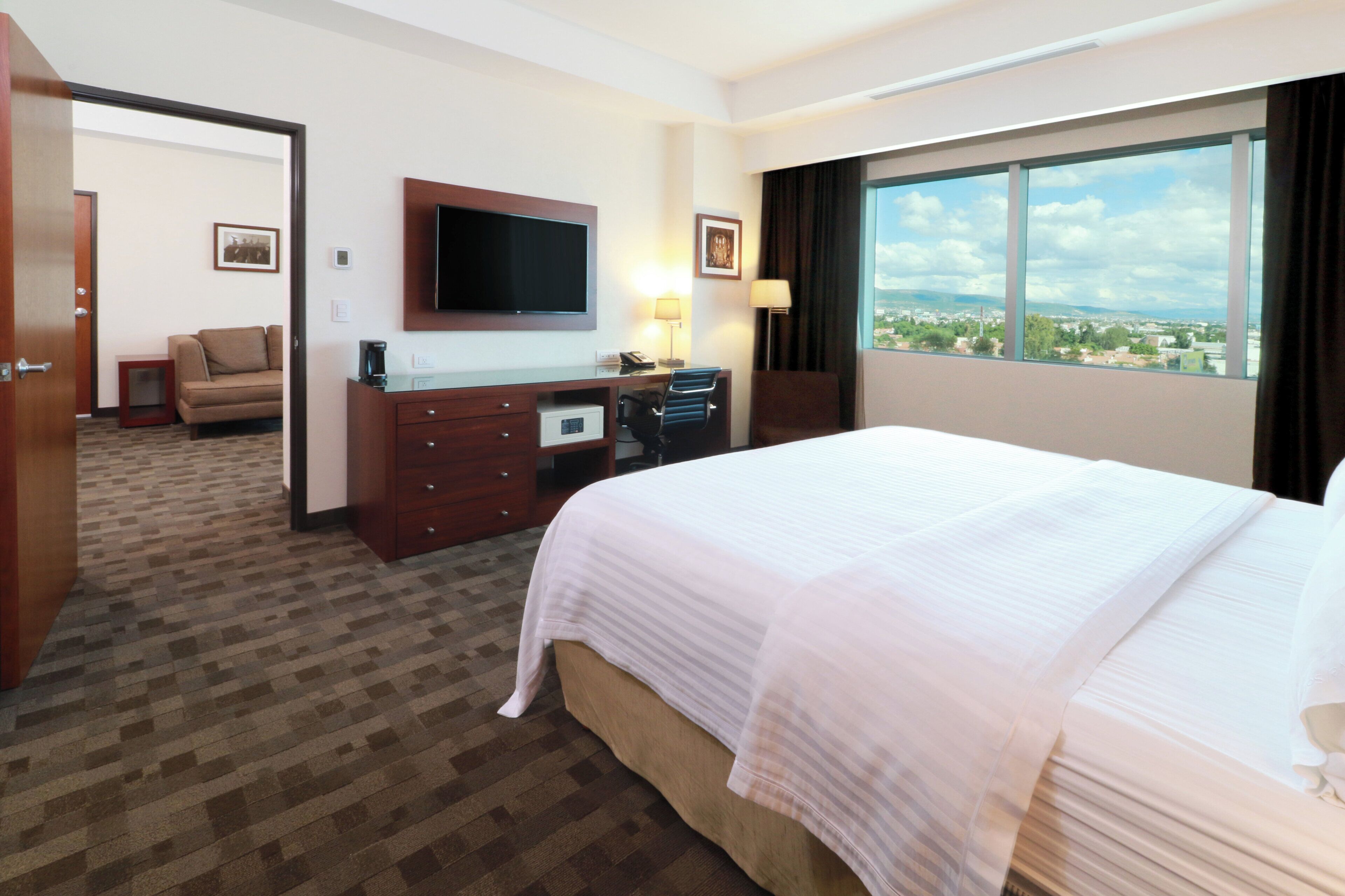 Habitación de hotel con cama, escritorio, televisión y vista a la ciudad.