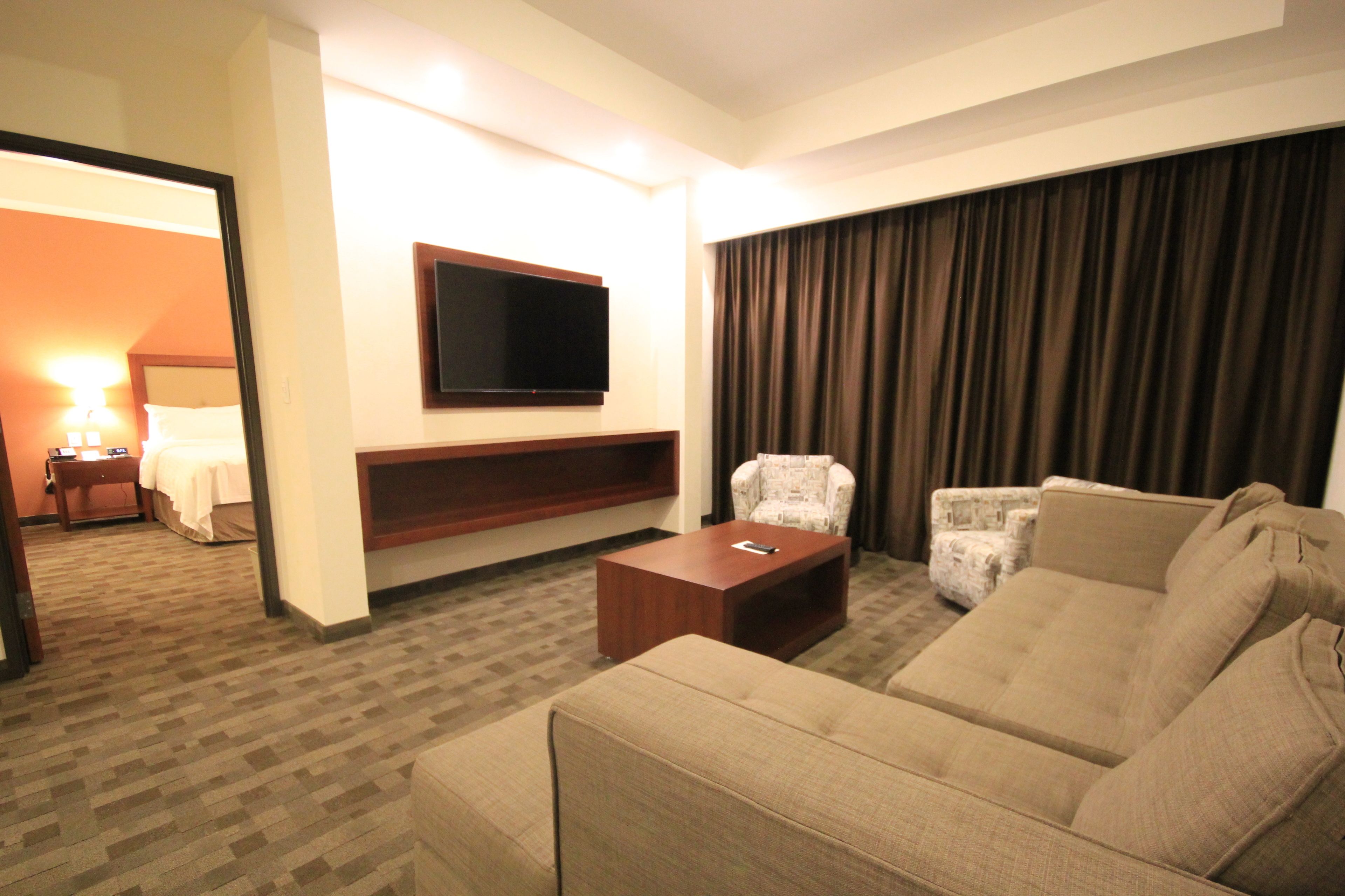 Habitación de hotel con un sofá grande, un televisor de pantalla plana montado en la pared, un sillón pequeño y una mesa de centro de madera.