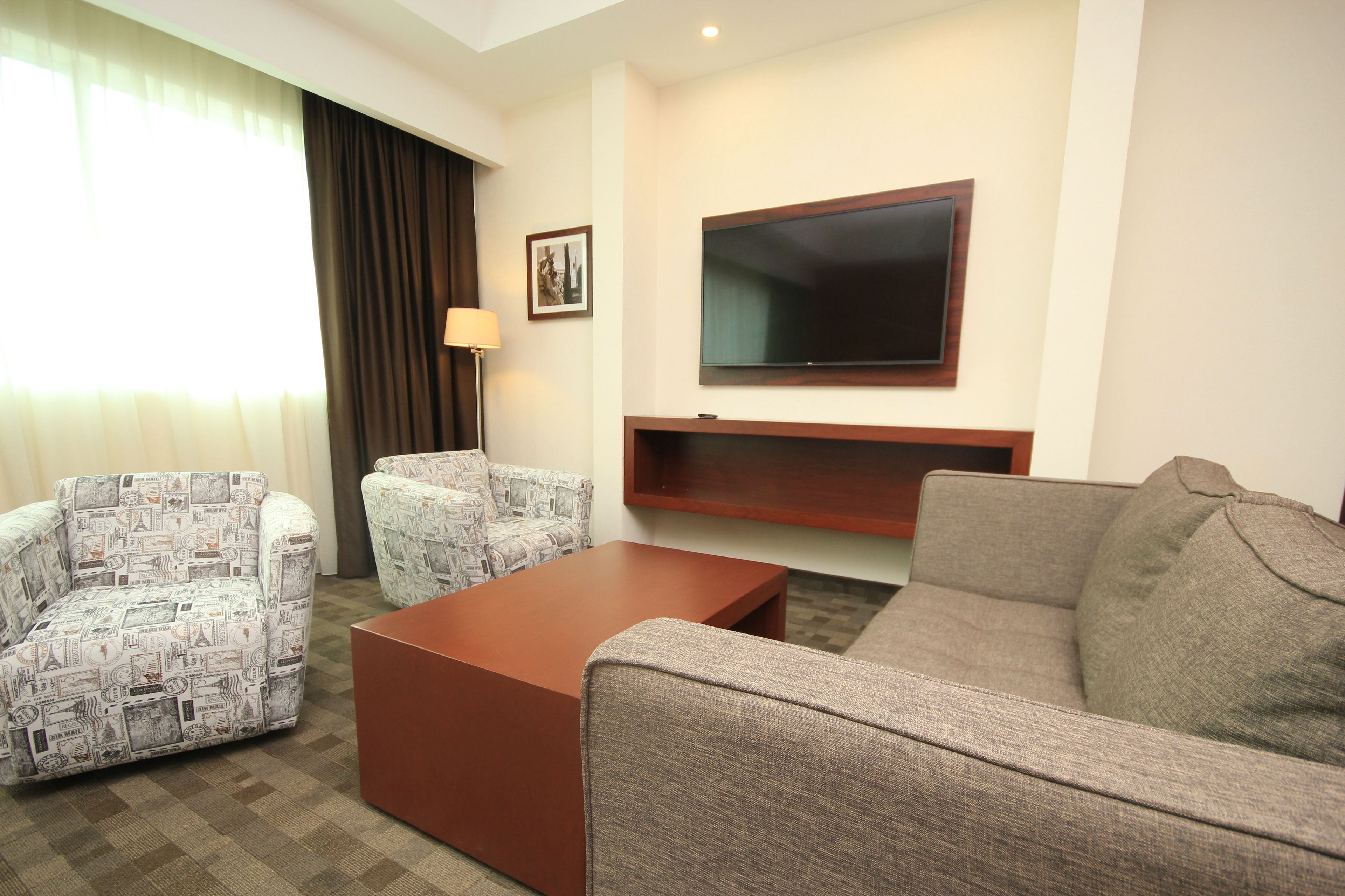 A living room with a brown sofa, two patterned armchairs, a coffee table, a flat-screen TV mounted on the wall, and a lamp.