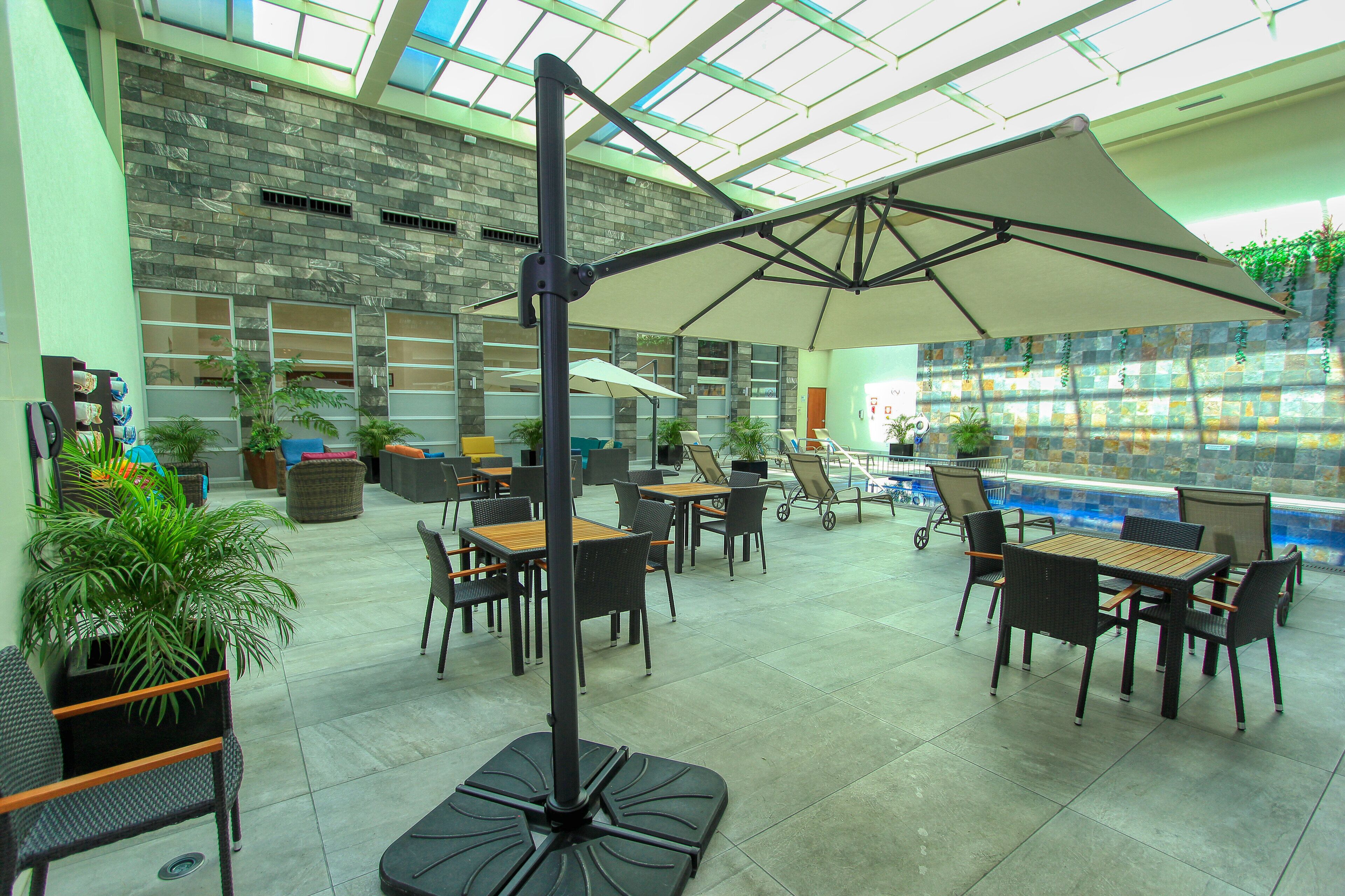 An indoor patio area with tables and chairs, a large umbrella, and a pool in the background.