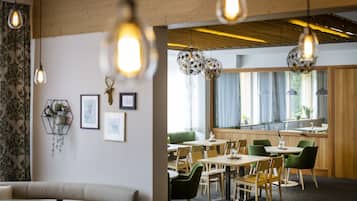 Un comedor moderno con mesas y sillas de madera, luces colgantes y vista a una cocina a través de un amplio ventanal.
