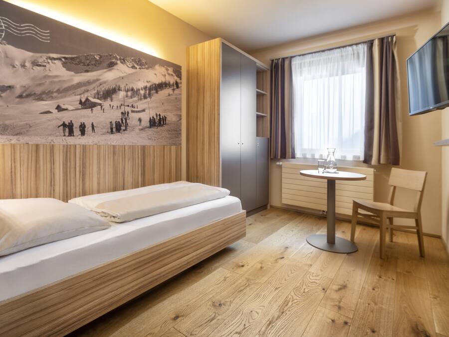 A modern hotel room with a bed, a small table, a chair, a TV, and a large wall-mounted picture.