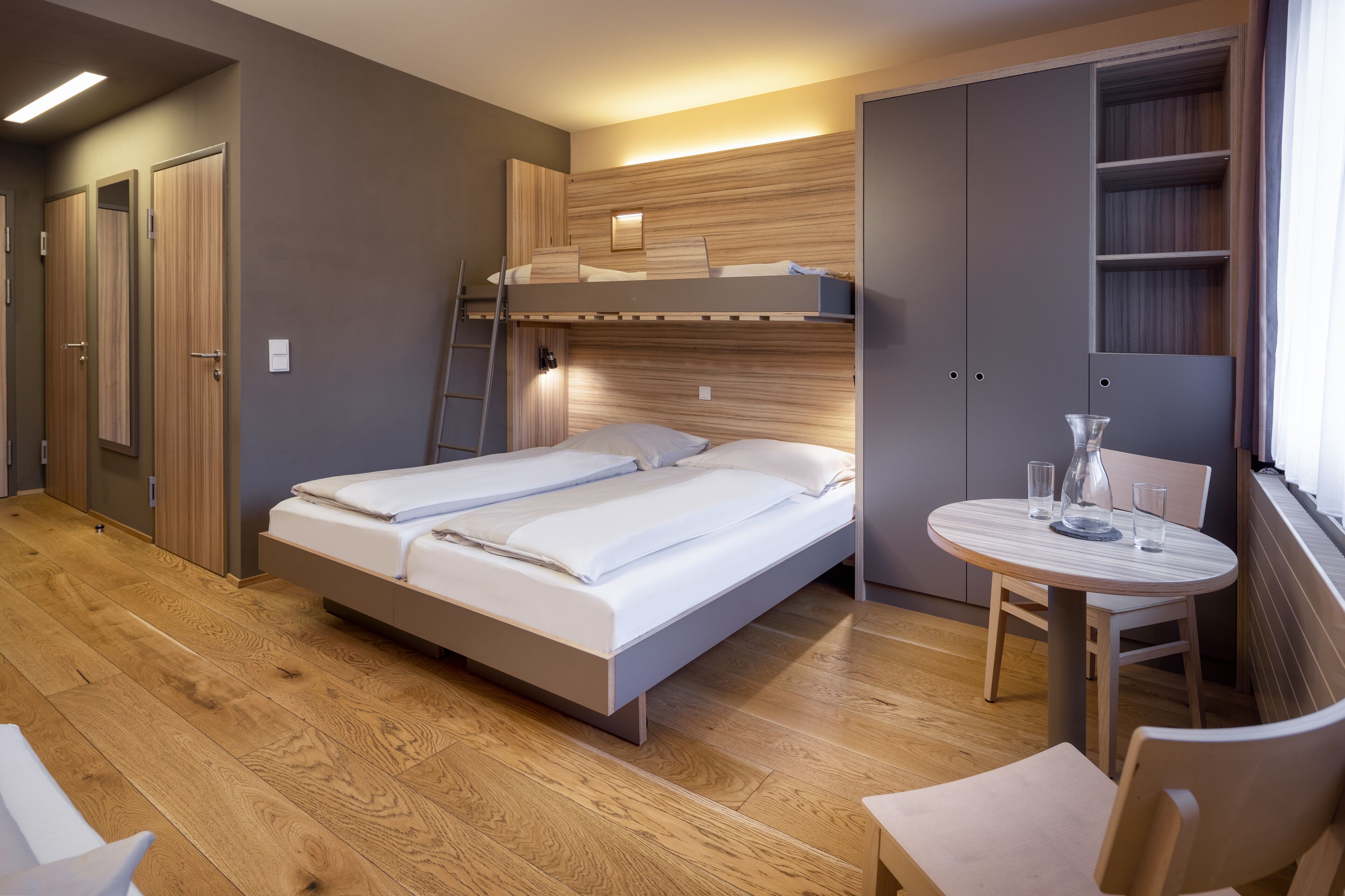 A modern hotel room with a bunk bed, a round wooden table, and built-in shelving.