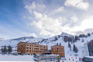 Un edificio de varios pisos con fachada de madera, situado en una zona montañosa nevada con pistas de esquí al fondo.