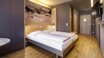 A modern bedroom with a wooden headboard, a bed with white linens, a mounted TV, and a mountain landscape mural on the wall.