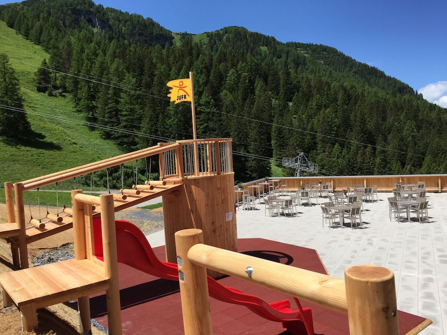 Un parque infantil de madera con tobogán y zona de escalada, rodeado por una terraza de madera con mesas y sillas, frente a un paisaje de colinas verdes y cielo despejado.