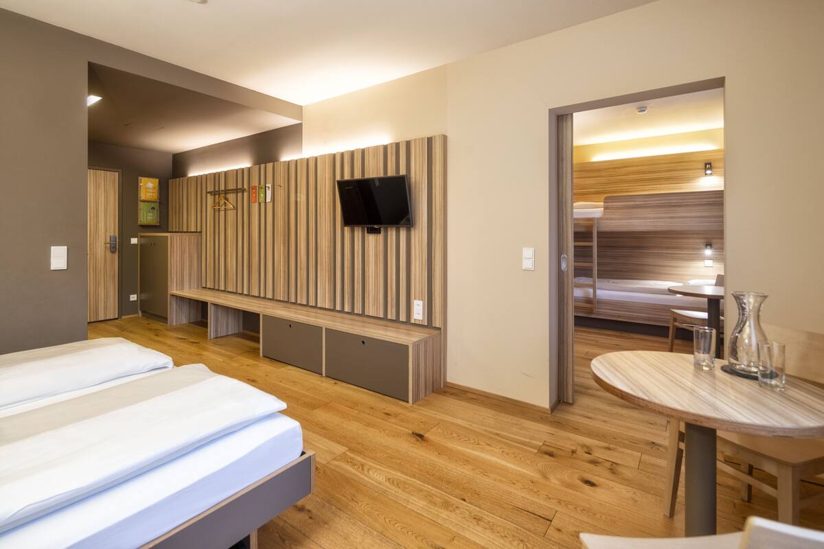 Une chambre d’hôtel moderne avec deux lits, un téléviseur fixé sur un mur recouvert de panneaux en bois, une petite table ronde avec des verres et un banc.