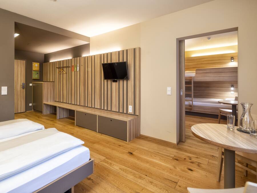 Une chambre d’hôtel moderne avec deux lits, un téléviseur fixé sur un mur recouvert de panneaux en bois, une petite table ronde avec des verres et un banc.
