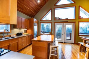 A modern kitchen with wooden cabinets, a central island, and large windows offering views of trees. - Stoke Cabin by Revelstoke Vacations (Revelstoke)