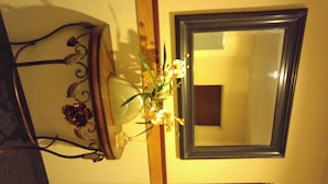 Un espejo con marco oscuro en una pared, una lámpara con pantalla blanca y un arreglo floral.