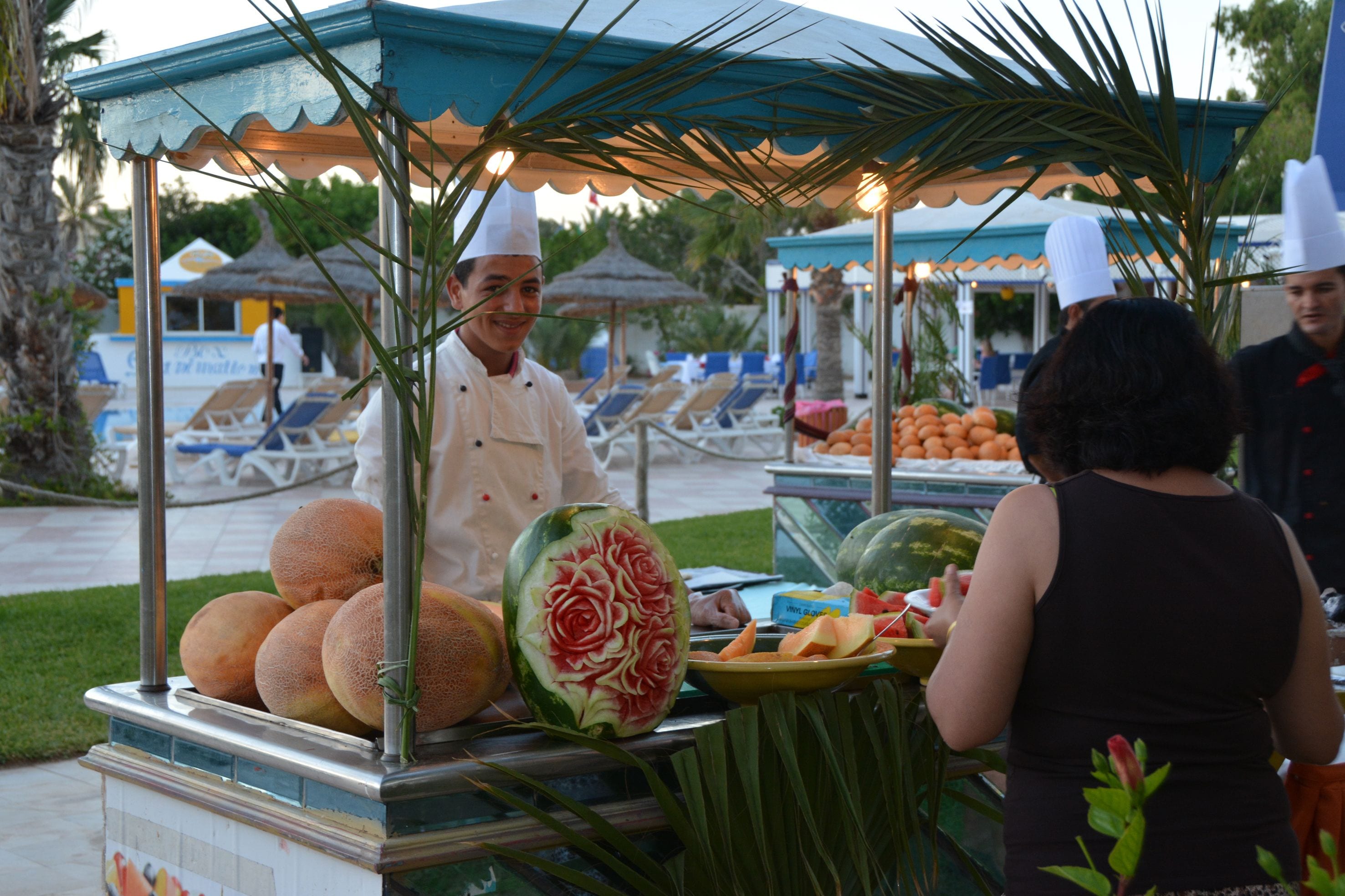 A food stall with a variety of fruits and vegetables, including a watermelon with a rose design, and a chef wearing a white hat.