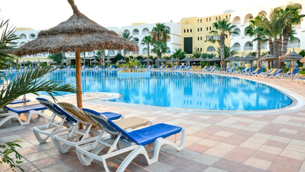 Une piscine entourée de chaises longues et de parasols, bordée de palmiers et d’un bâtiment d’hôtel doté de balcons.