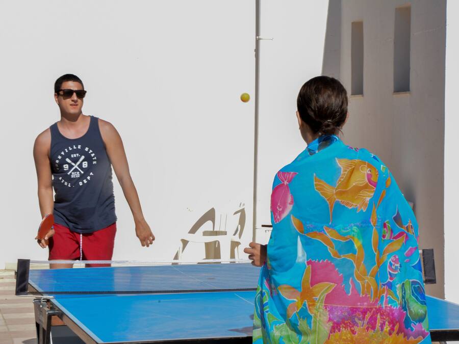 Dos personas jugando ping pong en una cancha al aire libre con una pared blanca y una mesa azul al fondo.