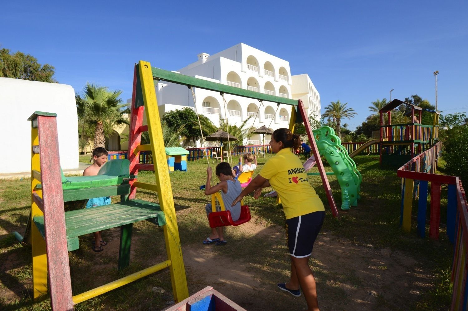 A playground with colorful equipment, including swings and slides, and a building with arched windows in the background.