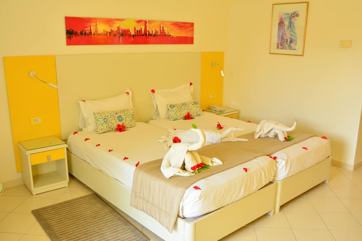A neatly made bed with white linens and decorative pillows, a bedside table with a lamp, and a framed cityscape painting on the wall.