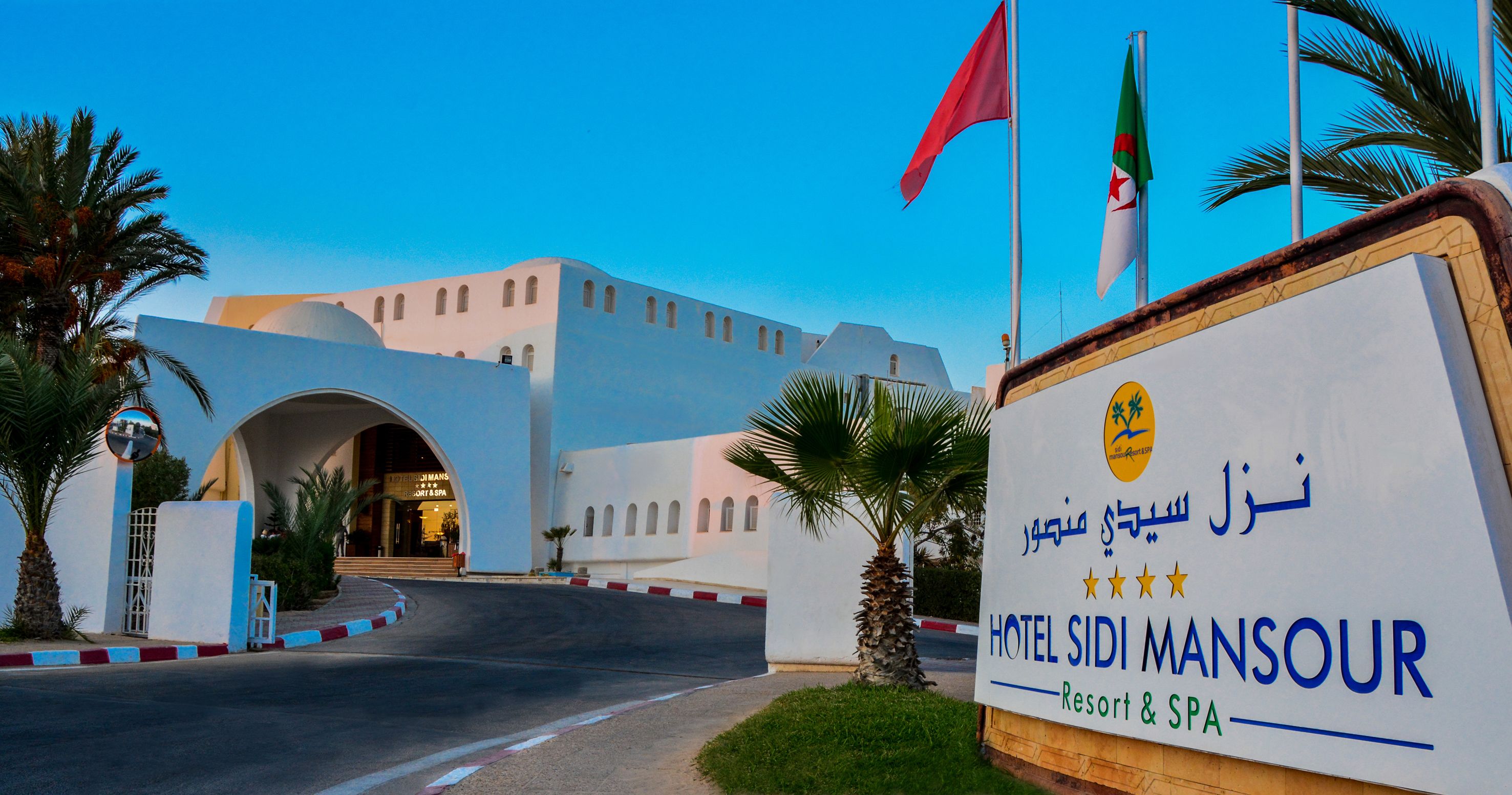 The entrance to Hotel Sidi Mansour, featuring a prominent sign with the hotel's name and logo, a clear view of the building's architecture, and a palm tree-lined pathway leading to the entrance.