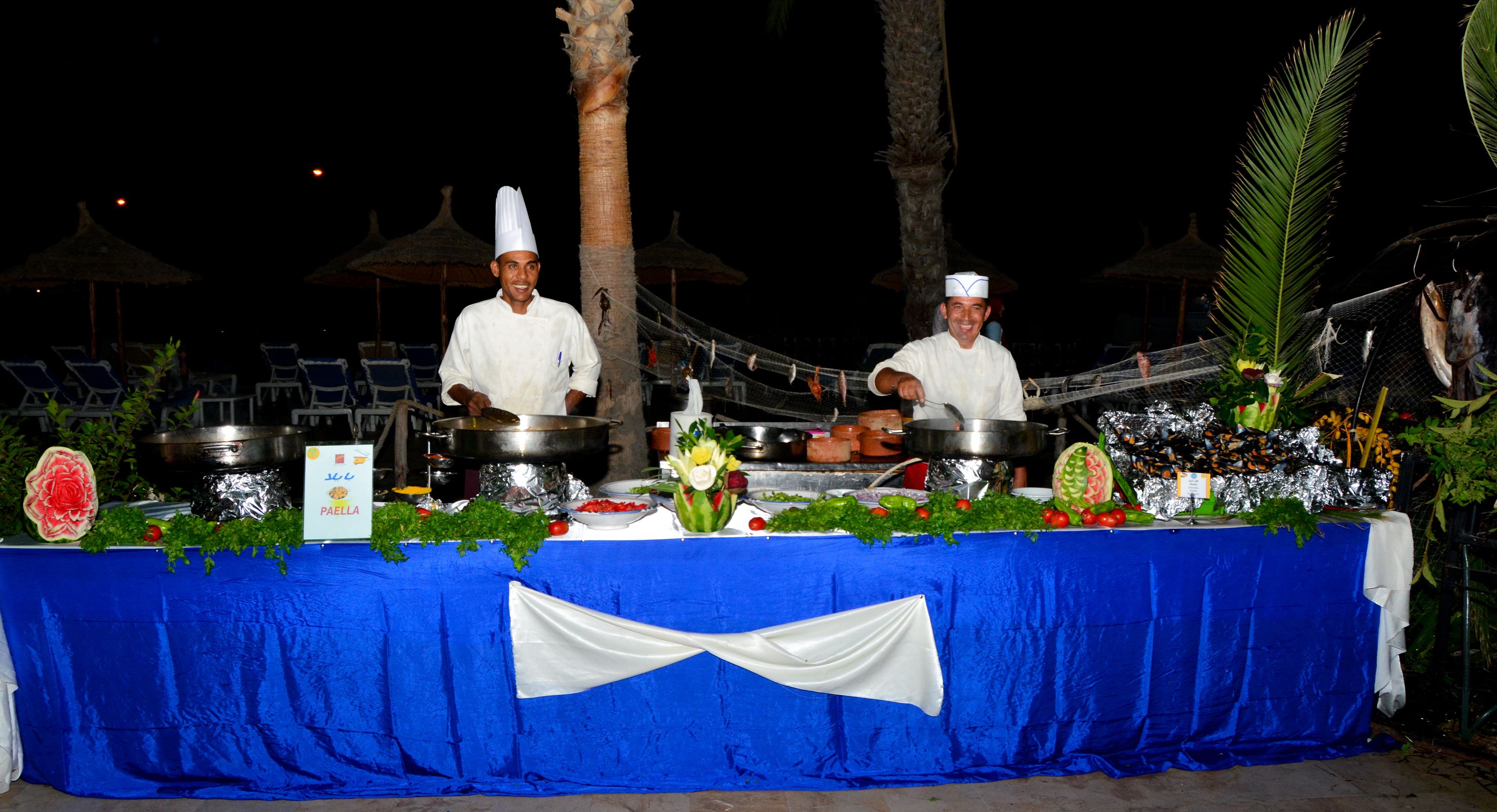 A buffet table with chefs, covered with a blue tablecloth and white drapes, and decorated with fresh vegetables and flowers.