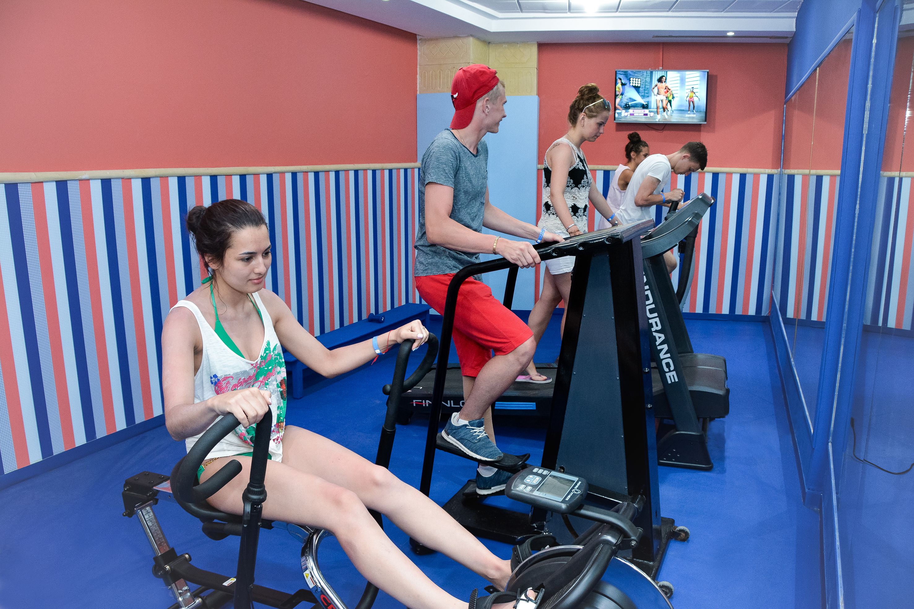 A gym with treadmills and stationary bikes, striped walls, and a television displaying fitness content.