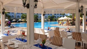 Un espace de restauration extérieur abrité, avec des tables disposées pour deux personnes, offrant une vue sur une piscine et un bâtiment orné d’arcades.