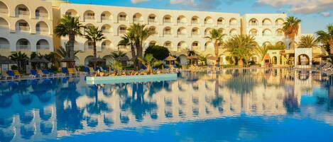 A large pool with clear blue water, surrounded by a hotel with multiple balconies and palm trees.