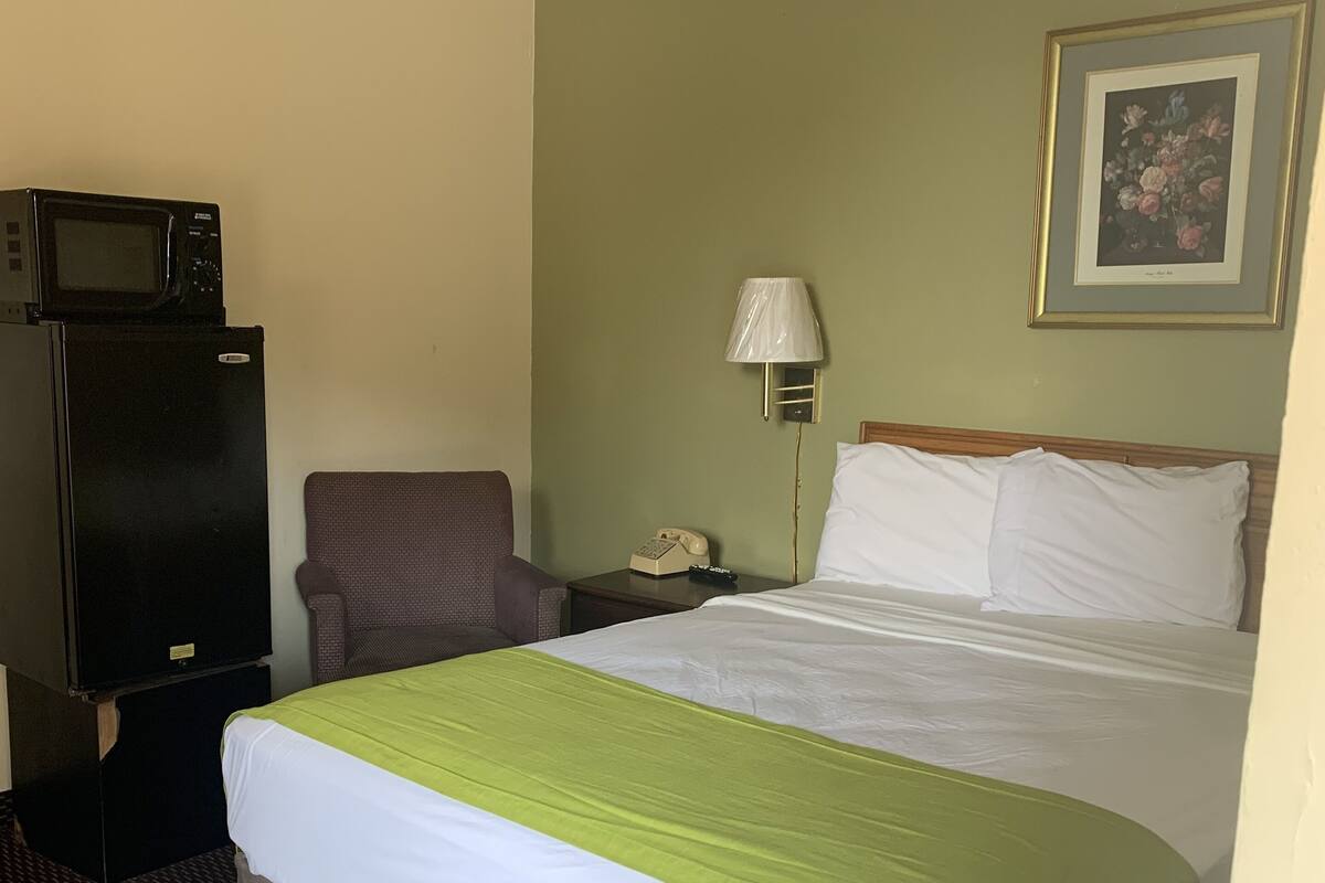 A hotel room with a bed, a chair, a desk with a lamp, a telephone, and a microwave on a refrigerator.