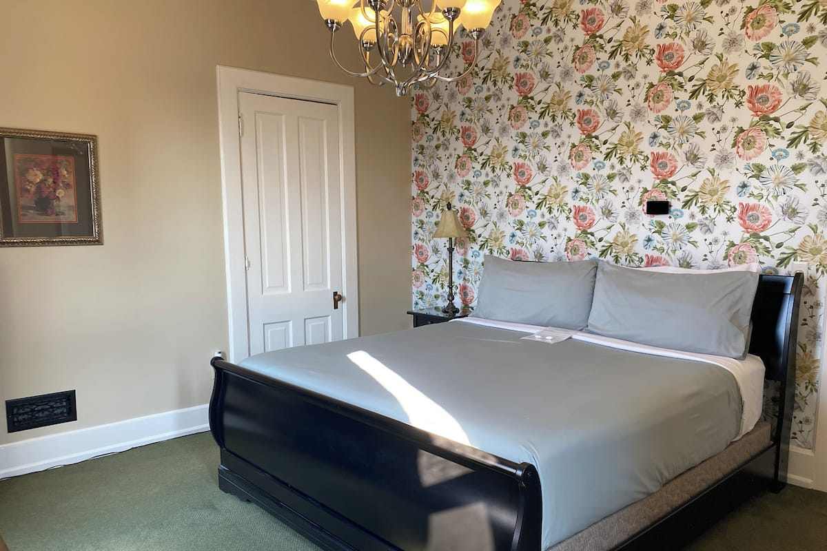 A bedroom with a bed, a door, a wall with floral wallpaper, and a chandelier.