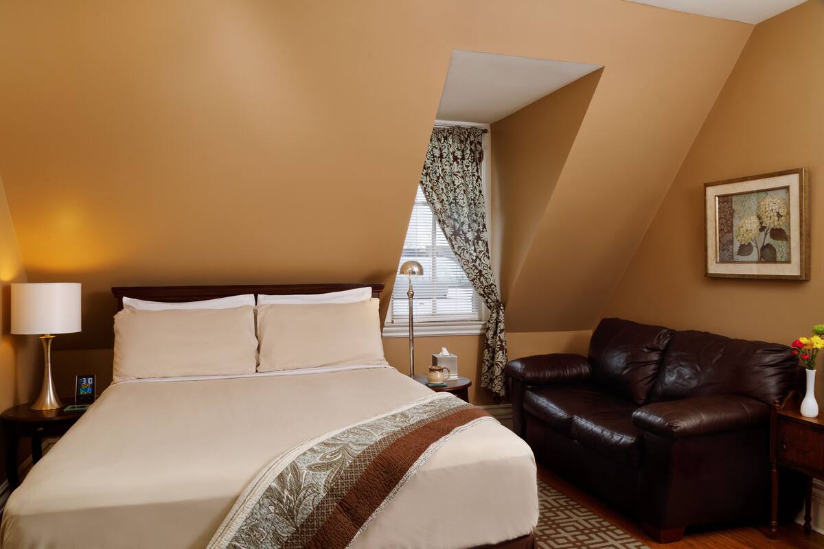 A hotel room with a large bed, a leather sofa, a bedside table with a lamp, a clock, and a window with curtains.