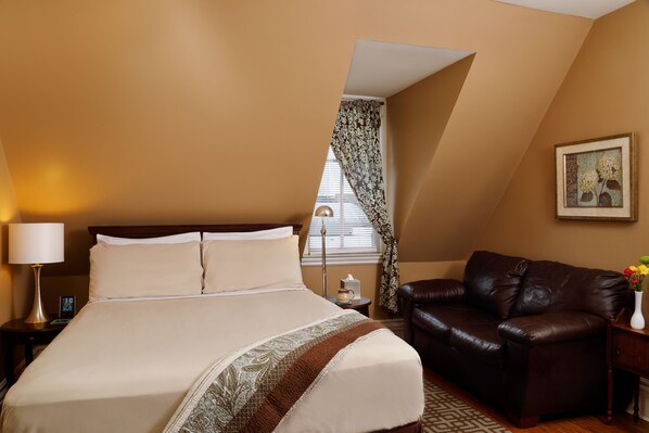 Quarto de hotel com uma cama grande, um sofá de couro, uma mesa de cabeceira com abajur, um relógio e uma janela com cortinas.