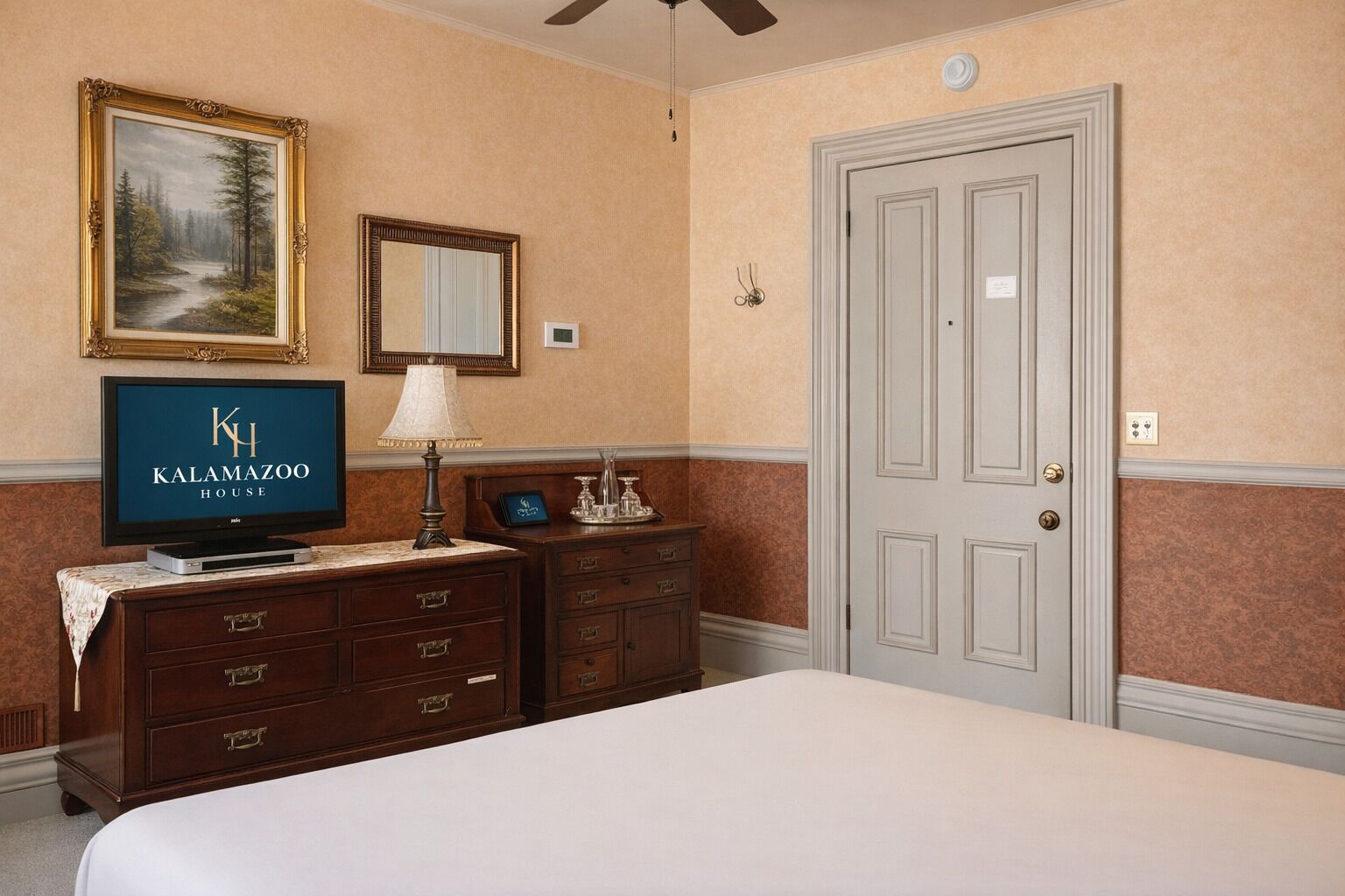 Une chambre d’hôtel avec un lit, un téléviseur diffusant l’émission « Kalamazoo House », une commode, un miroir et une porte.