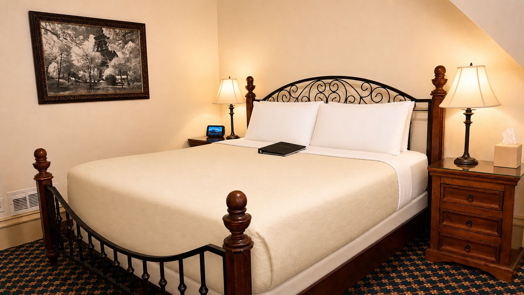 A neatly made bed with a wooden headboard, two bedside lamps, a bedside table with a lamp, and a framed picture on the wall.