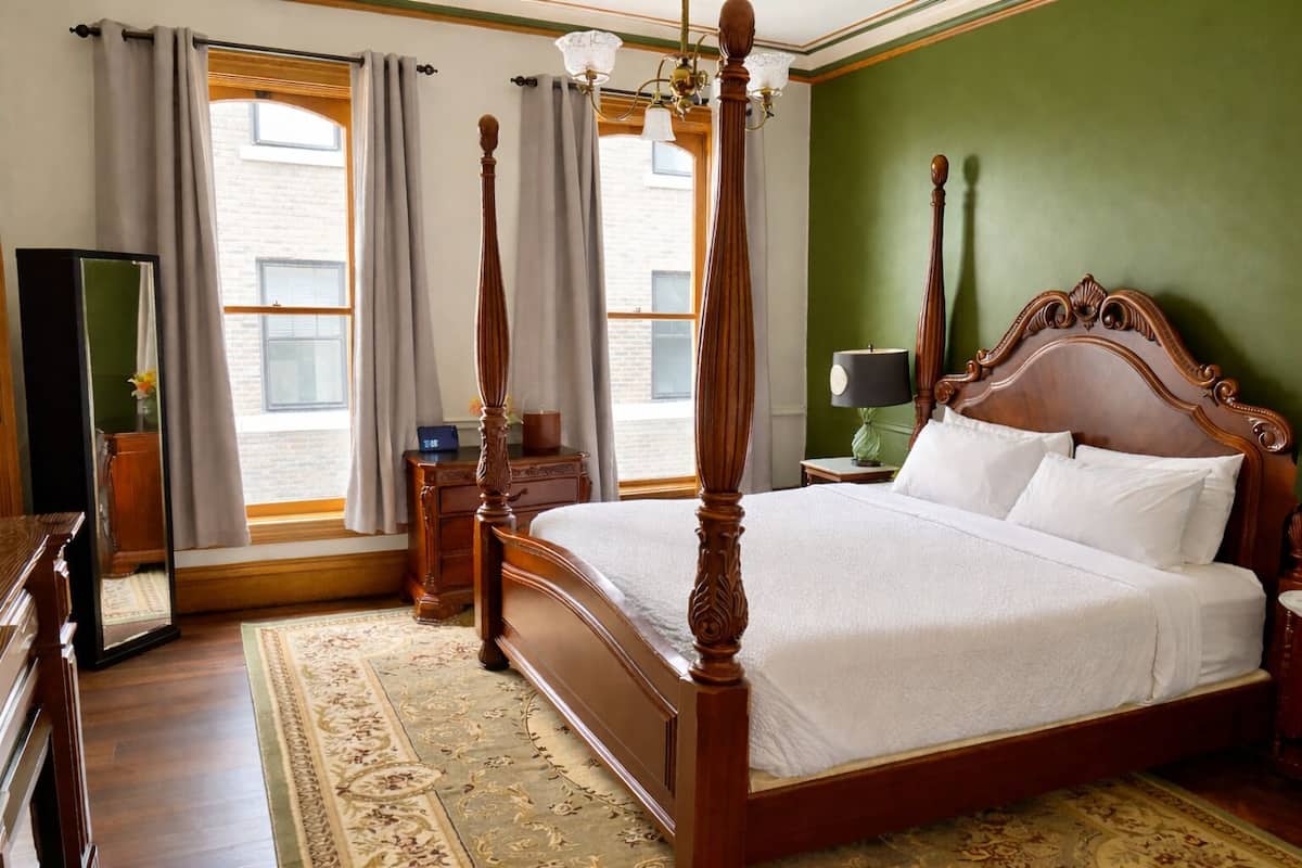 A four-poster bed with a wooden headboard, a bedside table with a lamp, a dresser, and a large mirror.