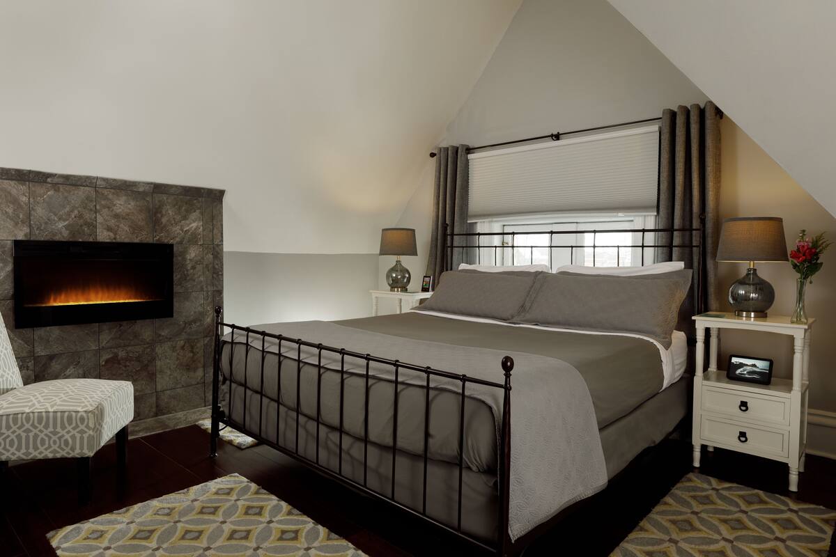 A bedroom with a bed, a TV mounted on the wall, a nightstand with a lamp, and a fireplace.