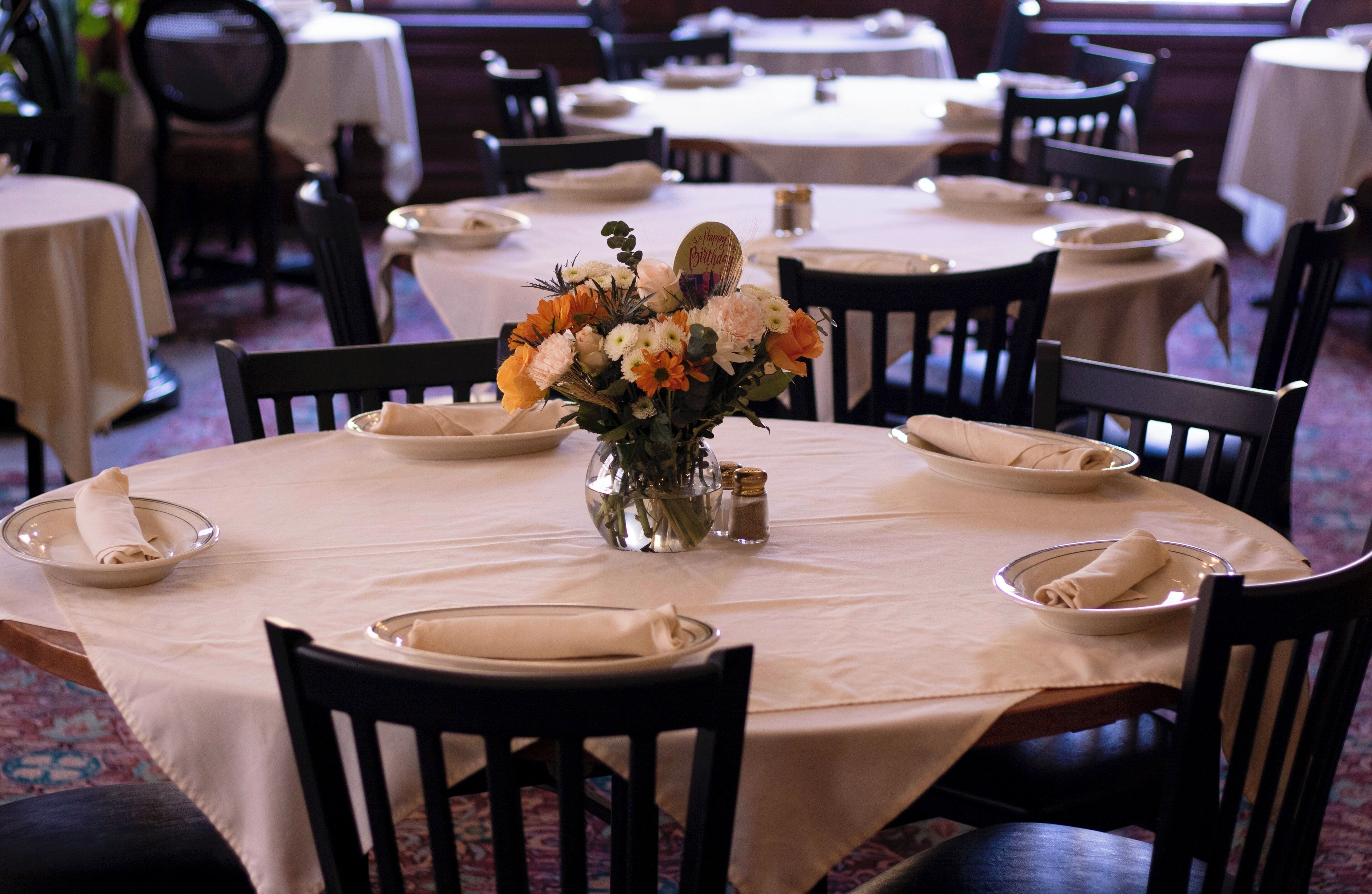 Un comedor con mesas redondas, cada una con mantel blanco, platos y servilletas dobladas. Un jarrón con flores se encuentra en el centro de una mesa.