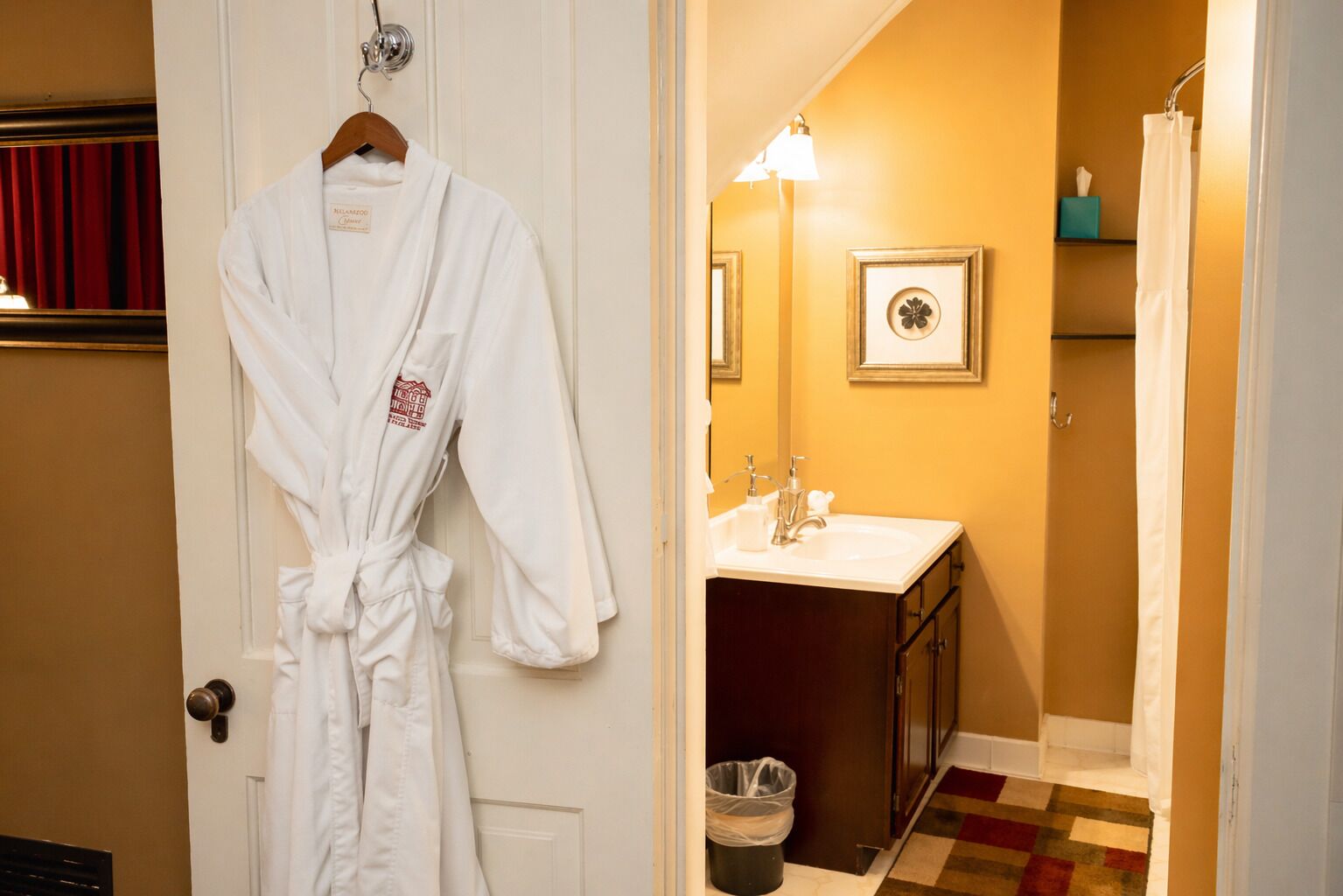 Habitación de hotel con una bata blanca colgada en la puerta, un baño con lavamanos y espejo, y una alfombra estampada.