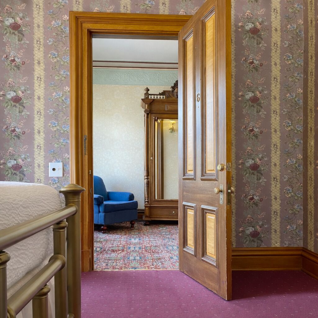 Una habitación con una cama, un sillón azul, un armario de madera y una alfombra estampada.