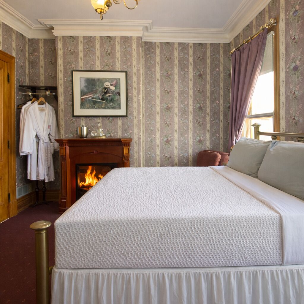 A bedroom with a large bed, a fireplace, a framed picture on the wall, and a window with curtains.